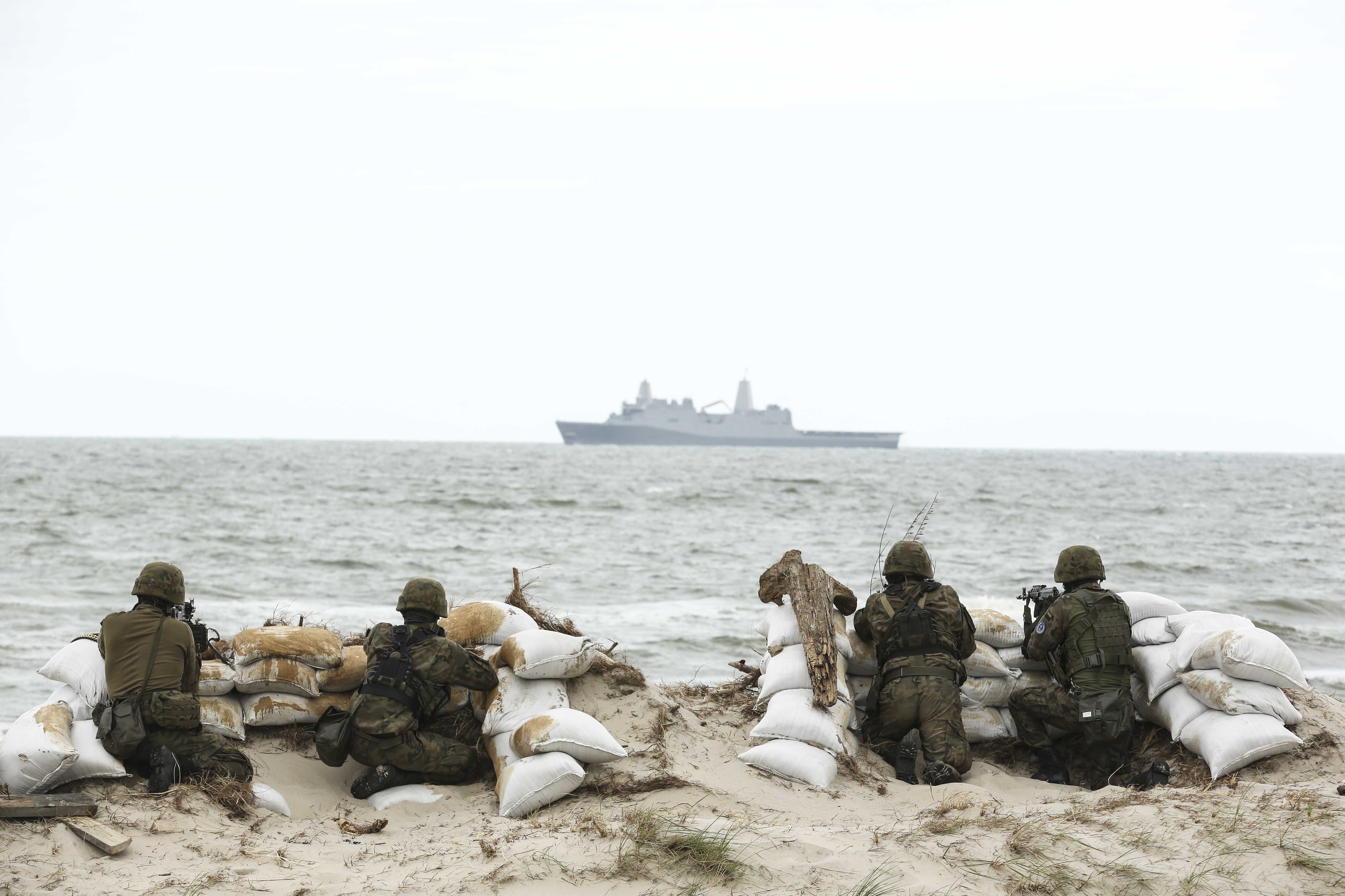 Soldater der gemmer sig bag sandsække og kigger ud over havet, hvor der er et stort skib
